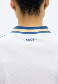 Dos de la nuque d'une personne portant une chemise blanche avec un col rayé bleu et jaune et "italia" brodé en bleu sous le col.