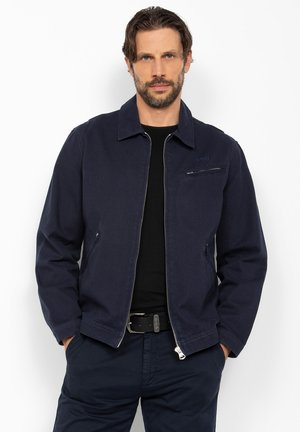 Homme barbu portant une veste bleu marine déroulée sur une chemise noire, debout les mains dans les poches devant un fond blanc.