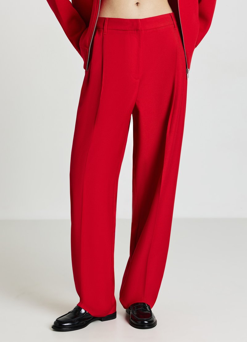 Pantalons taille haute rouges en tissu lisse et fluide avec une coupe large. Présente des plis droits et une coupe ajustée. Associés à des chaussures noires.