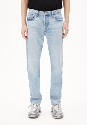 Lichtblauwe denim jeans met een rechte pijp, vijf zakken, zichtbare stiksels en een subtiele vervagede textuur. Gecombineerd met grijze sneakers.
