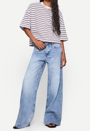 Femme portant un t-shirt rayé oversize, un jean bleu clair à jambes larges et des sandales noires, debout devant un fond blanc uni.