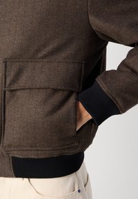 Bruine herringbone bomberjack met geribbelde zwarte manchetten en zoom, voorzien van zijzakken en een textuur voor extra dimensie.