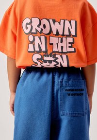 Enfant portant un t-shirt orange avec le texte « Grown in the Sun » et un graphique de soleil, assorti d'un pantalon bleu avec une poche arrière étiquetée « American Vintage ».