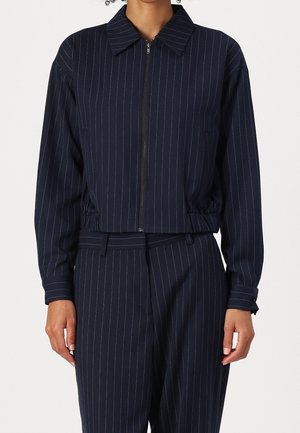 Femme portant une veste bleu marine à fines rayures avec une fermeture éclair frontale et un pantalon assorti, montrée de la nuque aux hanches, sur un fond uni.