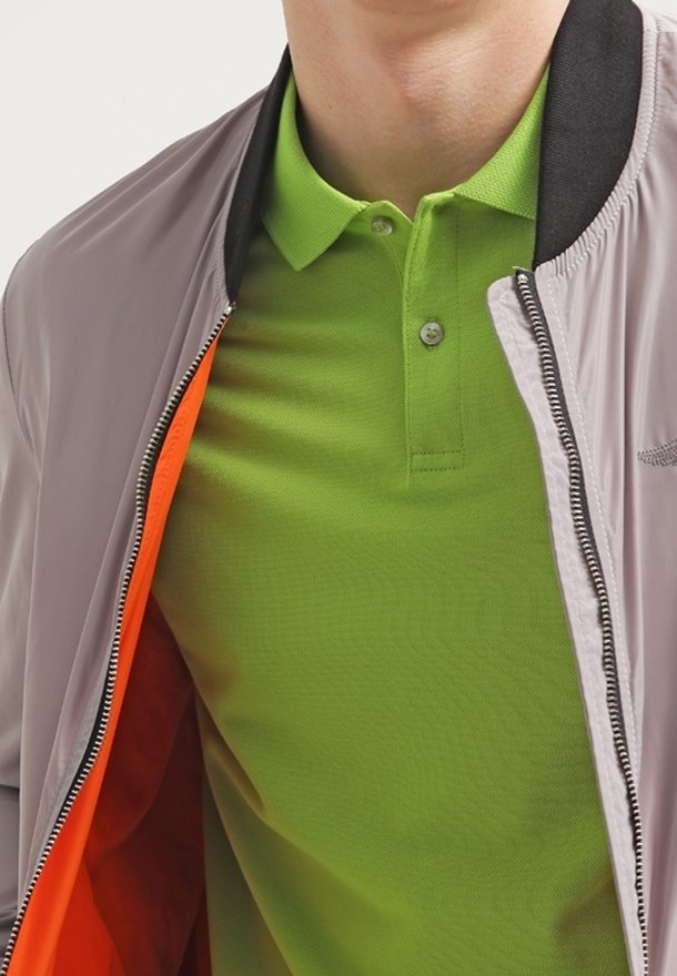 Veste bombardier grise avec doublure orange portée sur un polo vert vif. La veste présente un col noir et une texture lisse.