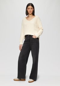 Strickpullover in Creme mit V-Ausschnitt, kombiniert mit lockeren schwarzen Jeans. Das Outfit wird durch tanfarbene und weiße Schuhe ergänzt.