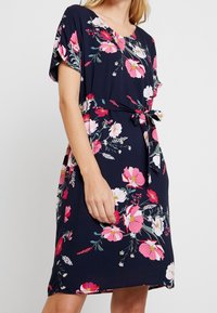 Blumenkleid mit dunkelblauem Hintergrund, kurzen Ärmeln, einem Bund mit Knoten und lebhaften rosa und weißen Blumenmustern. Glatte Stoffoberfläche.