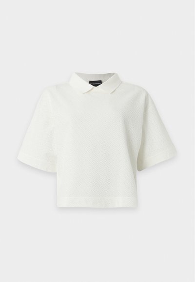 Top court blanc à manches courtes, texturé, avec un col pointu, une coupe ample et un motif chevron subtil, présenté sur un fond blanc uni.