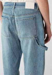 Lichtblauwe denim jeans gezien van de achterkant, met twee opgestikte zakken, een wit label op de tailleband en een zijlus voor het vastbinden van voorwerpen.