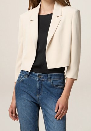 Kvinde iført kort cremefarvet blazer over sort top og blå jeans med små metalliske nitter, stående med afslappede arme.