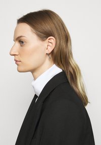 Silverfärgade hoops med en slät, polerad yta, mot en ljus bakgrund. Modellen bär en svart blazer och en vit polotröja.