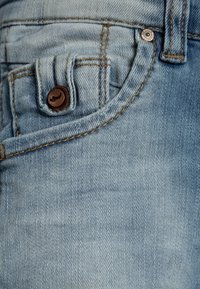 Jean en denim bleu clair avec un bouton marron, des coutures contrastées et un design de poche distinctif avec une texture subtile.