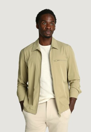 Uomo con una giacca beige con zip sopra una camicia bianca e pantaloni chiari, in piedi con le mani nelle tasche, che guarda leggermente di lato.