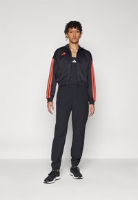 Črno športno oblačilo, ki vključuje kratko jakno z oranžnimi črtami, majico brez rokavov in zožene hlače. Zasnovano za udobje in gibljivost.