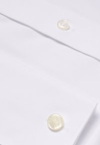 Chemise en coton blanc avec deux boutons crème gravés du logo, texture lisse et col structuré. Détail de design ajusté visible.