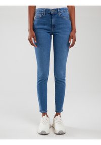 Cilvēks, kurš valkā pieguļošas zilas džinsa bikses un baltus sporta apavus, stāv pret vienkāršu gaišu fonu.