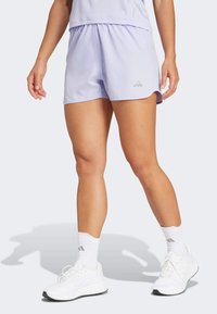 Pantalones cortos atléticos de color púrpura claro con cintura fruncida, que presentan un logo reflectante sutil y una textura suave. Combinados con zapatos y calcetas blancas.