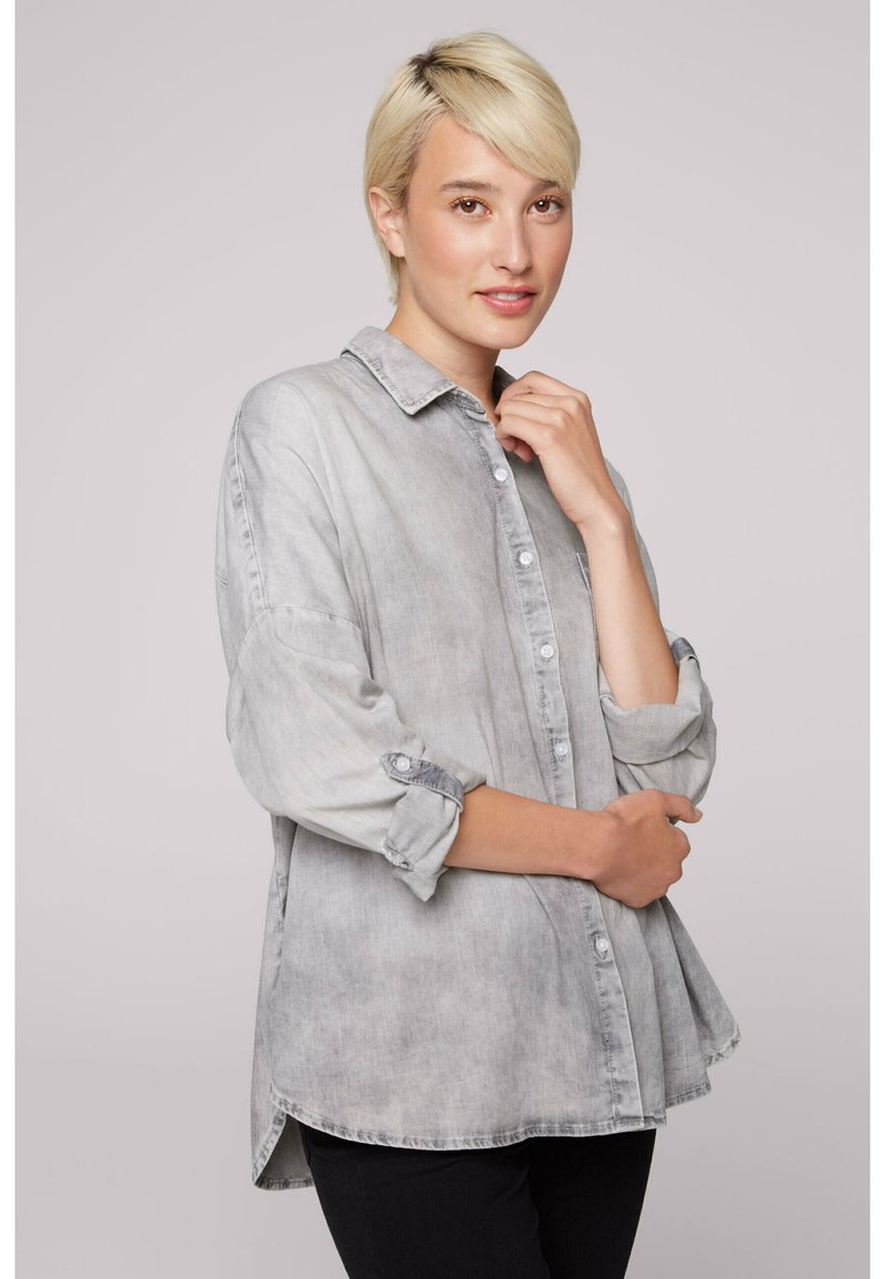 Light gray denim shirt with button front, point collar, and rolled sleeves. Features a relaxed fit and a curved hem. No visible patterns.