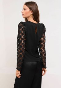 Juoda palaidinė su gėlių nėriniais ilgomis rankovėmis, turinti atvartą nugaroje ir tekstūruotą detalę. Lygus audinys kūno dalyje kontrastuoja su rankovėmis.