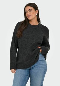 Frau mit langen dunklen Haaren, die silberne Ohrringe, einen dunkelgrauen gerippten Pullover und hellblaue Jeans trägt, posiert mit einer Hand im Pulloverbund.