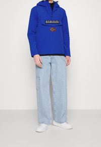 Osoba ve modré bundě s kapucí a přední kapsou, s nášivkou vlajky, ve světle modrých širokých džínách a bílých teniskách na jednobarevném pozadí.