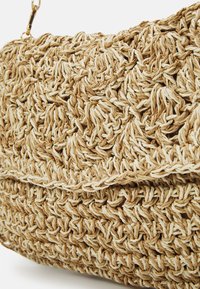 Beige vävd väska gjord av naturliga fibrer, med en strukturerad form och en texturerad yta med flätade mönster. Guldiga detaljer.
