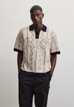 Homme portant un polo en maille beige à manches courtes avec col et poignets noirs, rayures florales verticales et texte "PEGADOR" sur la poitrine.
