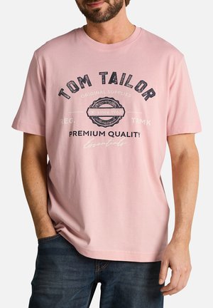 Homme portant un t-shirt Tom Tailor rose clair avec le texte et l'emblème « Premium Quality Essentials », associé à un jean bleu foncé.