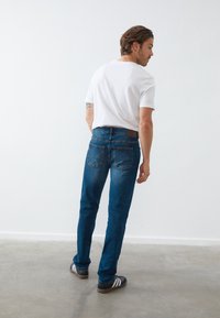 Hombre con camiseta blanca, jeans azules y zapatillas negras Adidas, de pie en un interior sobre un suelo de concreto frente a una pared blanca lisa.