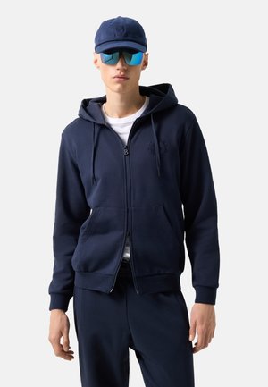 Man wearing navy zip-up hoodie, matching pants, white T-shirt, blue cap, and reflective sports sunglasses, standing against plain background.