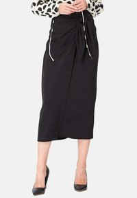 Jupe midi noire en tissu doux, avec un détail de nœud torsadé à l'avant et des liens sur les côtés, associée à des escarpins noirs à bout pointu.