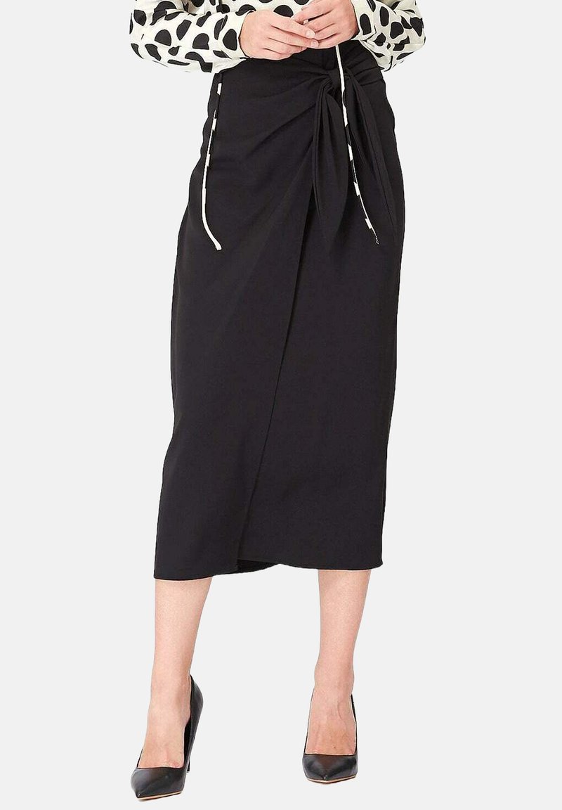 Jupe midi noire en tissu doux, avec un détail de nœud torsadé à l'avant et des liens sur les côtés, associée à des escarpins noirs à bout pointu.