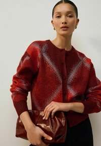 Červený pletený cardigan s geometrickým vzorem, který má kulatý výstřih, zapínání na knoflíky a žebrované manžety. Modelka drží vínovou psaníčko.