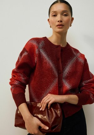Rode gebreide cardigan met een geometrisch patroon, voorzien van een ronde halslijn, knoopsluiting en ribgebreide manchetten. Het model houdt een kastanjebruine clutch tas vast.