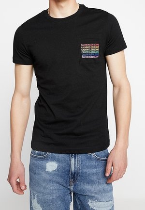 Homme portant un t-shirt noir à manches courtes avec le texte multicolore « Calvin Klein Jeans » sur la poche poitrine et un jean bleu déchiré.