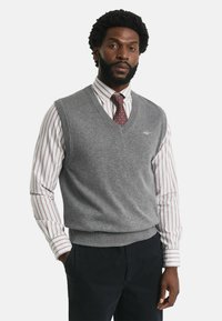 Grijze gebreide sweatervest met een V-halsontwerp, gedragen over een wit met rood gestreept shirt en gecombineerd met donkere broeken. Subtiel logo zichtbaar.