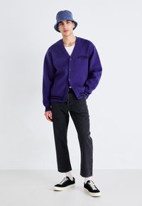 Cárdigan de punto morado con botones negros y rayas, llevado sobre una camiseta blanca. Combinado con jeans oscuros y zapatillas negras. Sombrero tipo cubo azul.