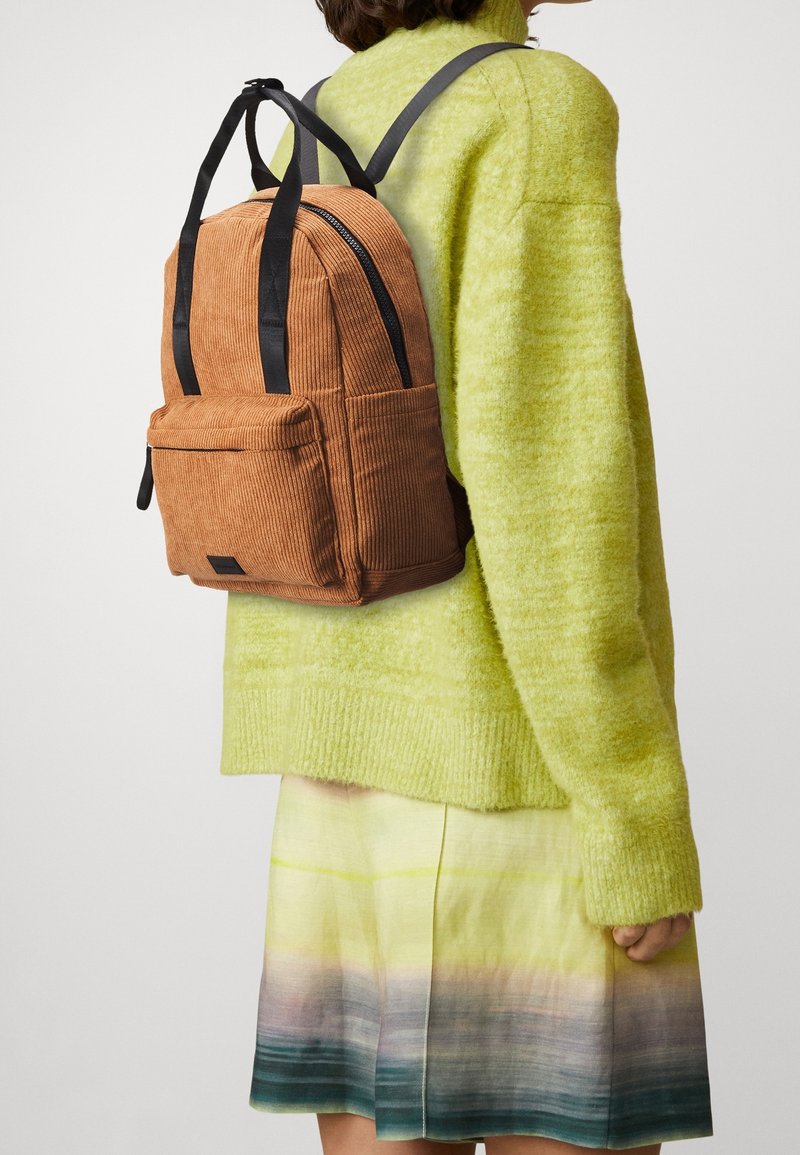 Femme portant un pull vert citron vert et une jupe multicolore avec un sac à dos en velours côtelé marron avec des bretelles noires dans son dos sur un fond uni.