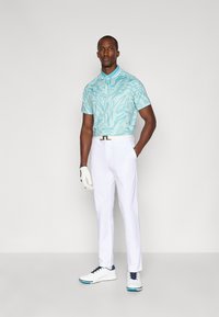 Polo de mangas curtas em teal com padrão abstrato, combinado com calças brancas e sapatos de golfe brancos com detalhes em azul-marinho. Segurando uma luva de golfe.