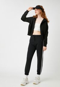 Sudadera corta negra con cremallera, top corto blanco de canalé, joggers negros con rayas laterales blancas y zapatillas deportivas blancas gruesas. Textura de tela suave.