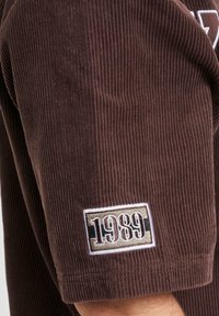 Camicia in corduroy marrone con maniche corte, tessuto testurizzato e un patch ricamato con il numero "1989" sulla manica.