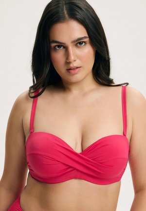 Młoda kobieta o ciemnych włosach, ubrana w jasno różowy top od bikini na gładkim jasnym tle, zwrócona przodem.