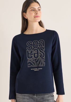 Frau trägt ein marineblaues Langarmshirt mit dem Text "sea tas" und "seaside ease moments" in Beige sowie abstrakten Wellenmustern auf der Vorderseite.