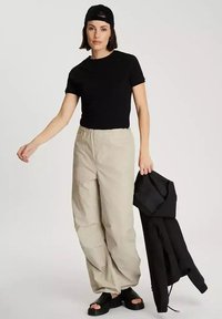 Czarna koszulka, beżowe szerokie spodnie, czapka w kolorze czarnym oraz czarna kurtka trzymana w ręku. Czarną parę sandałów dopełnia zestaw. Gładki materiał, casualowy styl.