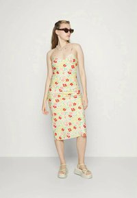 Vestido midi com padrão floral em amarelo pálido, apresentando flores vermelhas e rosas, alças finas e um silhueta ajustada. Combinado com sandálias de plataforma leves.