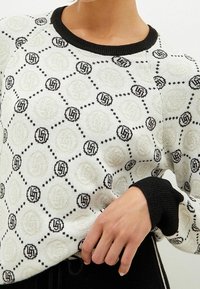 Strickpullover in Creme mit schwarzen Akzenten, der ein geometrisches Muster aus Kreisen und Sechsecken aufweist. Rippstrickbündchen in Schwarz an den Ärmeln und am Kragen.