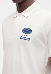 Personne portant un polo couleur crème avec "Passion Studios," quatre étoiles et "Hollister" logos brodés en bleu sur la poitrine.