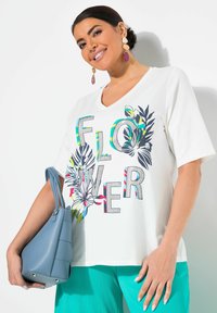 Camiseta blanca de cuello en V con gran texto "FLORES" y gráficos florales en verde, azul y rosa. Combinada con pantalones turquesa y un bolso azul.