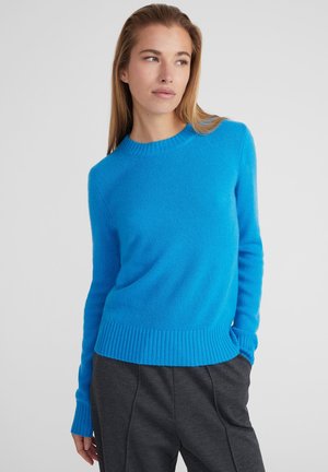Maglione lavorato a maglia blu brillante con scollatura rotonda, polsini e orlo a coste. Texture morbida, design aderente, indossato con pantaloni grigio scuro.