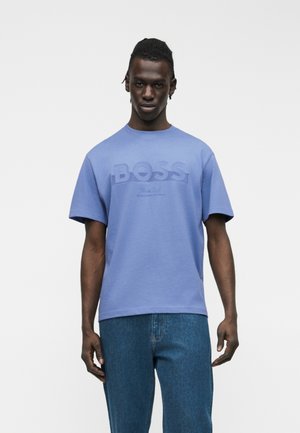 Férfi világoskék BOSS pólóban és kék farmernadrágban, egyszínű fehér háttér előtt áll, előre nézve, laza testtartásban.